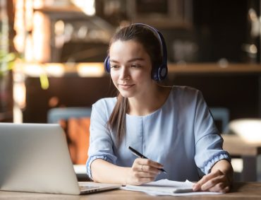 Focused,Woman,Wearing,Headphones,Using,Laptop,In,Cafe,,Writing,Notes,